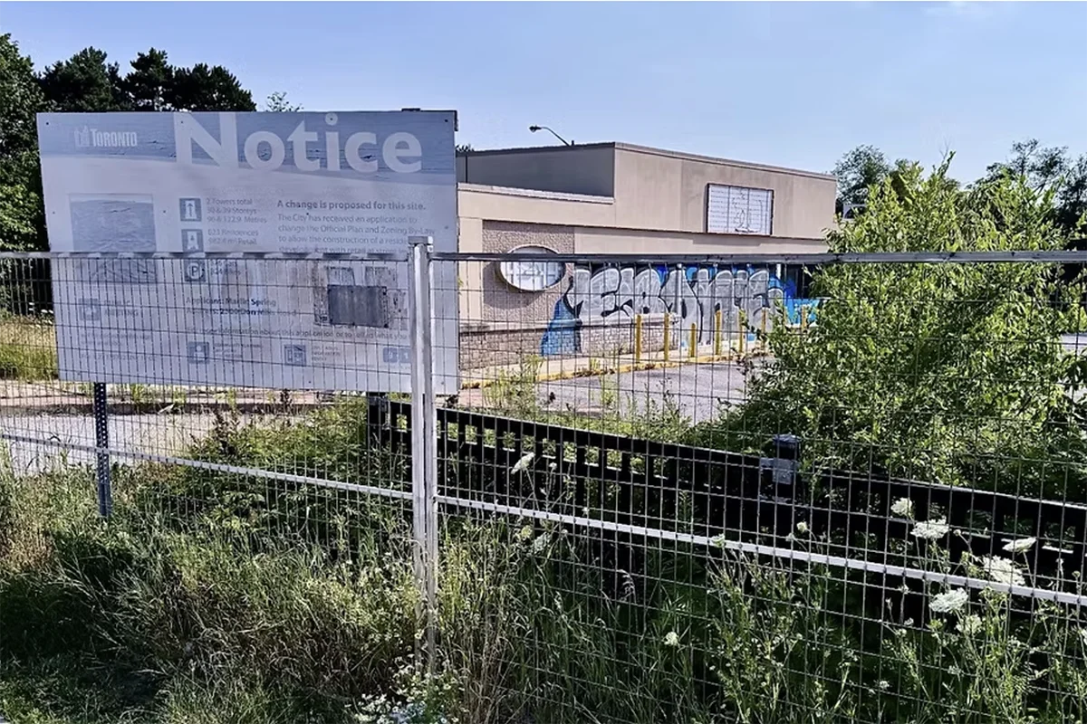 Boarded-up building surrounded by fencing and weeds, with faded development notice sign.